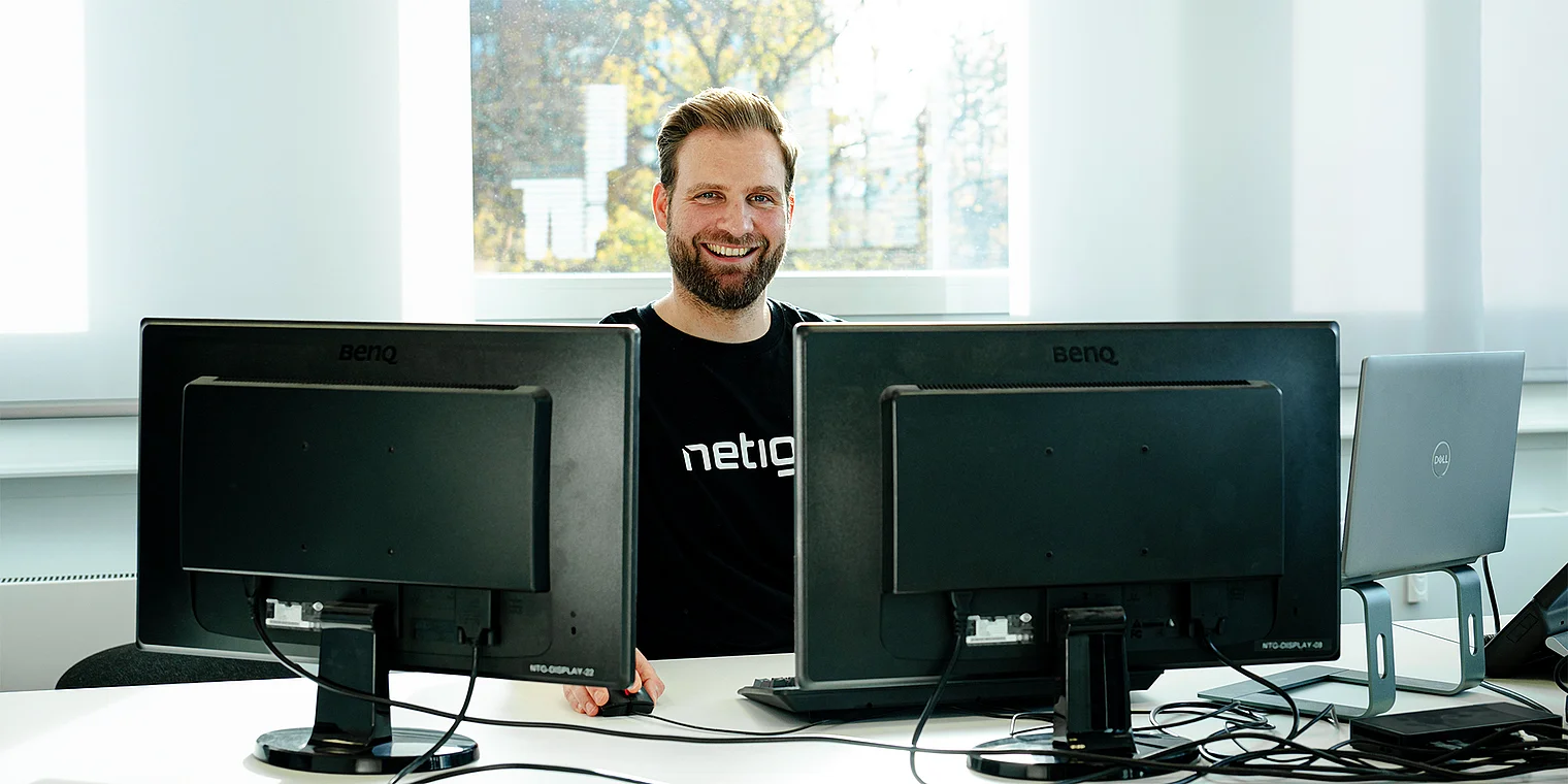 Ein lächelnder Mann sitzt an einem Schreibtisch mit zwei Monitoren und einem Laptop, umgeben von Kabeln und einem hellen Raum.