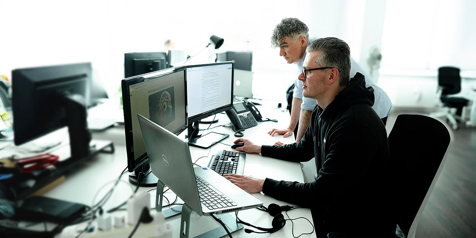 Walter und Kemal arbeiten fokussiert an einem digitalen Projekt und repräsentieren die AI-Optimierungs-Leistungen der Webagentur Netigo; der Hintergrund unterstreicht Themen wie künstliche Intelligenz, datenbasierte Analyse und technologische Innovation.