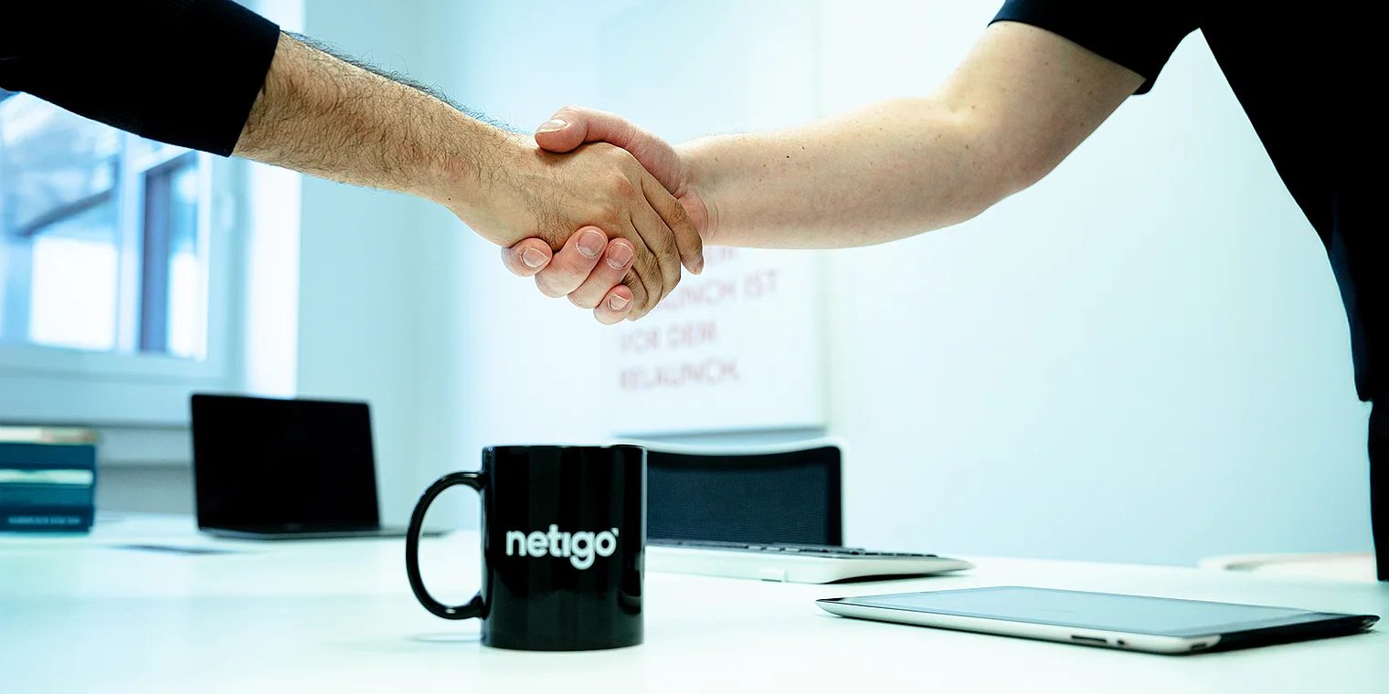 In einem hellen Büro umarmen sich zwei Hände in einem Handschlag, während ein Laptop und ein Tablet auf dem Tisch liegen.